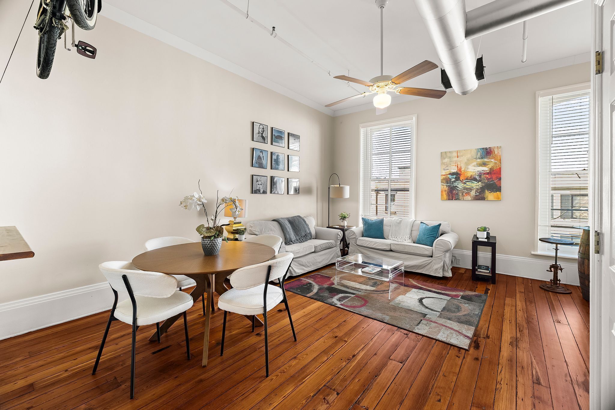 A spacious living room with hardwood flooring, a ceiling fan, a dining area with a round table and chairs, and a large window with blinds.