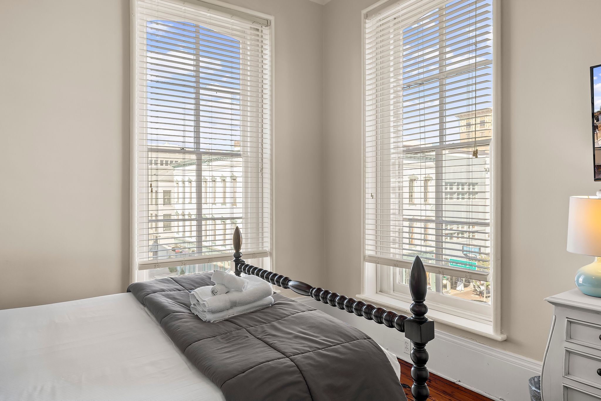 A bedroom with a bed, two windows with blinds, a bedside table with a lamp, and a view of buildings outside.