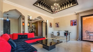 A modern living room with a black and red sofa, a glass coffee table, and a chandelier. There is a dining area with white chairs and a table, and a kitchen in the background.