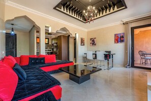 A modern living room with a black and red sofa, a glass coffee table, and a chandelier. There is a dining area with white chairs and a table, and a kitchen in the background. - River Palm Oasis, Pools, Netflix & Comfort (Marrakech)