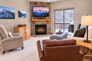 A living room with a fireplace, a television mounted on a stone wall, and a view of a snowy balcony.