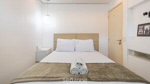 A neatly made bed with white linens, a brown throw blanket, and rolled towels on the bed.