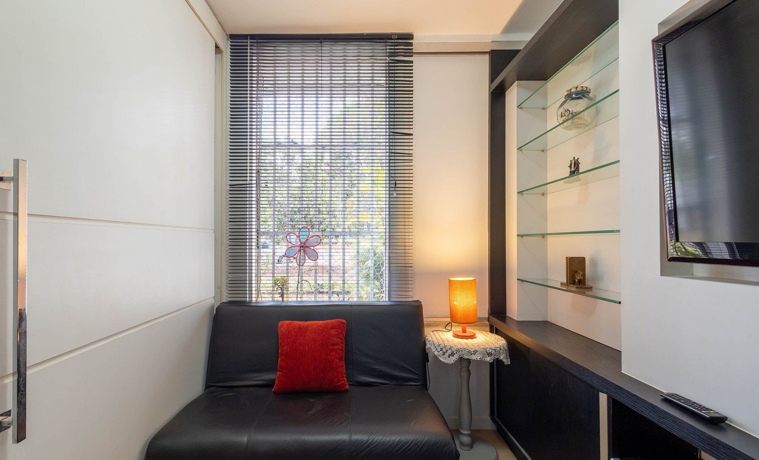 Een moderne woonkamer met een zwarte leren bank, een rode kussen, een kleine tafel met een lamp en een glazen plank met decoratieve objecten.