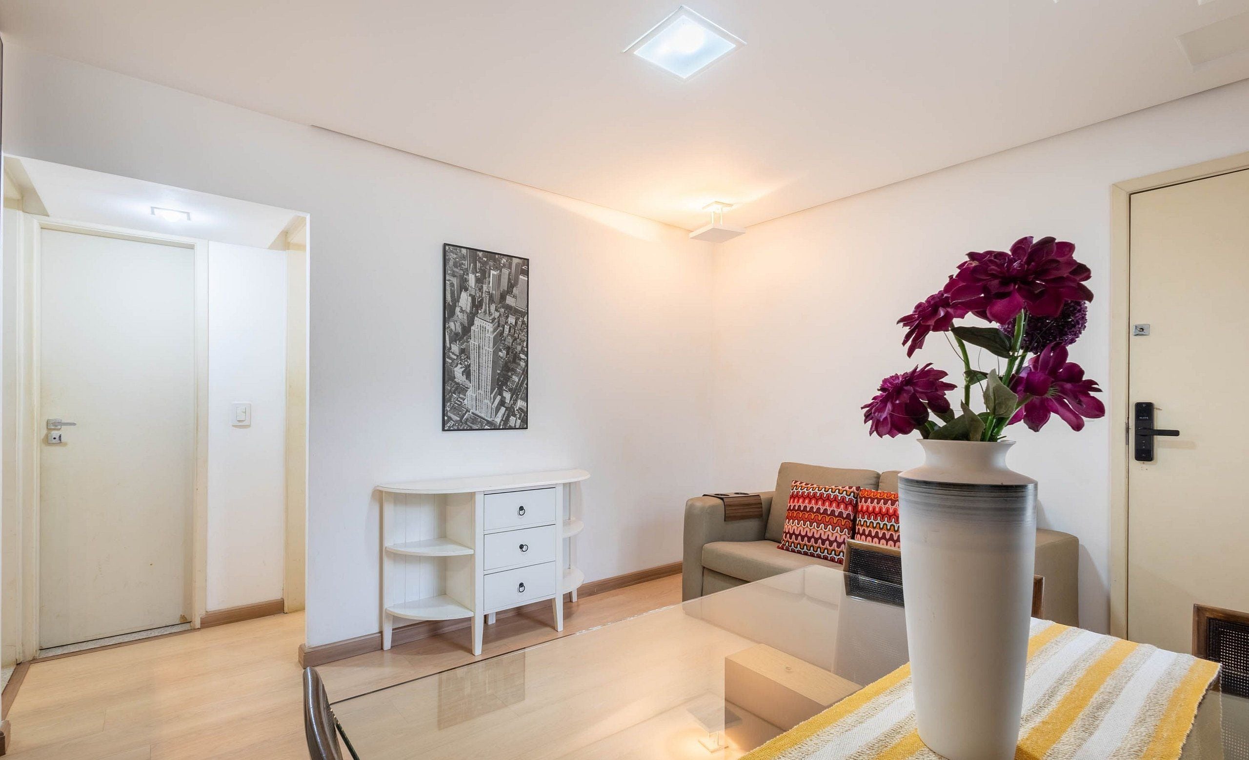 Un salon moderne avec une table basse en verre, un canapé blanc, un petit meuble blanc et un vase rempli de fleurs.