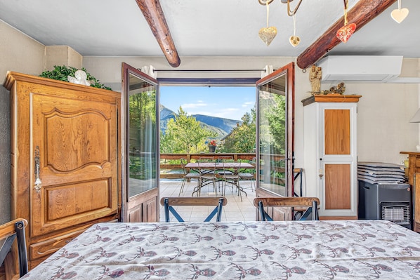 A room with a bed, wooden wardrobe, open door to a balcony with a table and chairs, and a view of mountains.