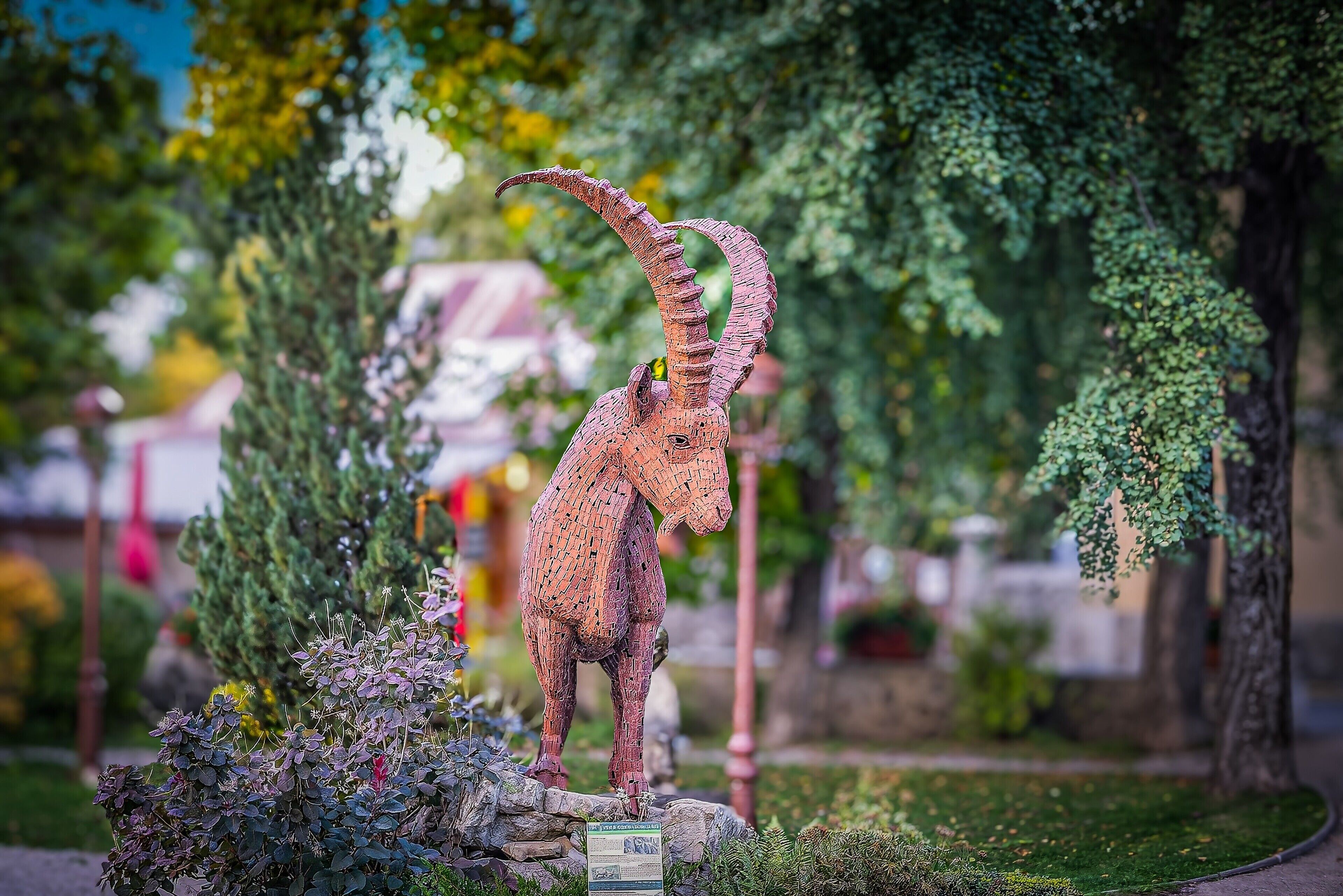 Una escultura de una criatura parecida a una jirafa, con cuello largo y cuernos curvados, rodeada de follaje y un pedestal de piedra.
