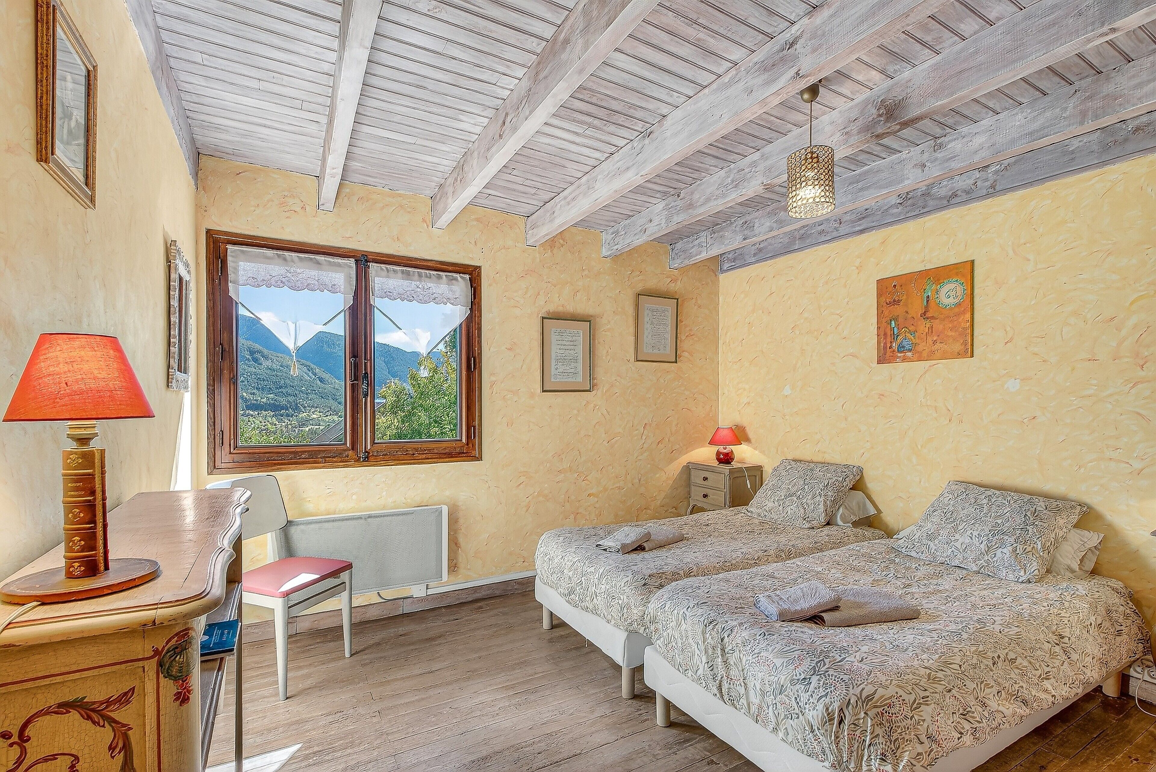 A bedroom with two beds, a desk with a lamp, a chair, a window with a view of mountains, and a ceiling with exposed beams.