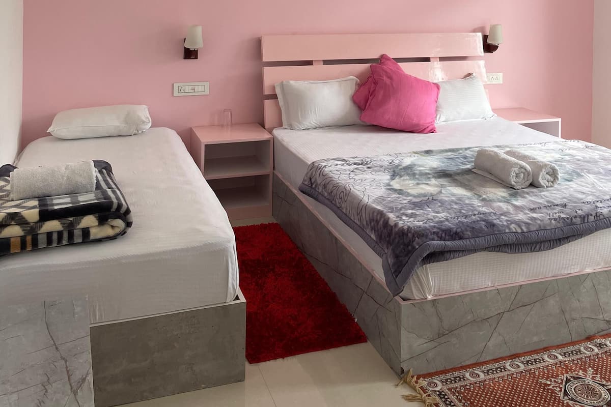 A hotel room with a pink wall, a bed with a white and gray patterned bedspread, a red rug, and a ceiling fan.