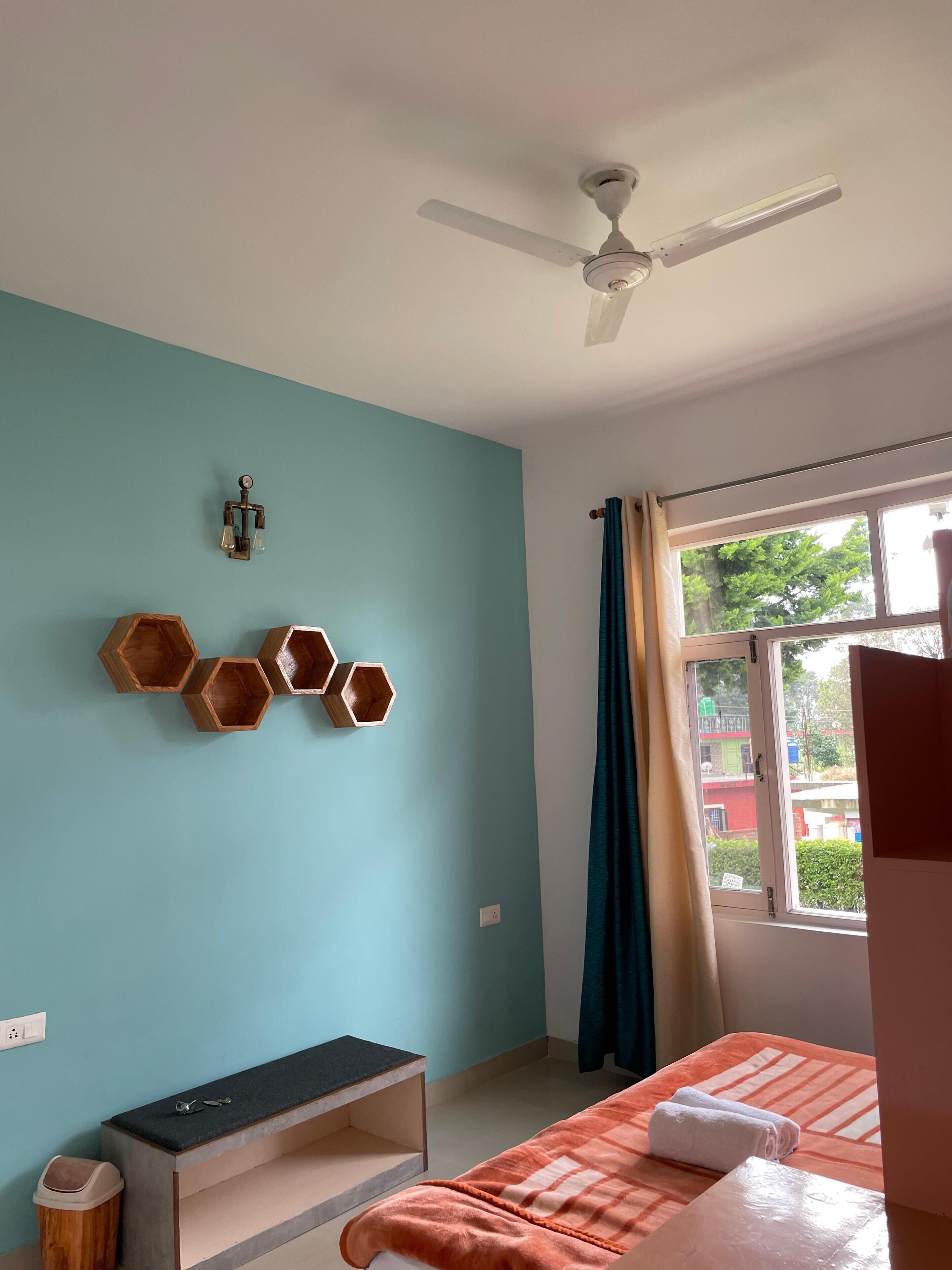 A room with a teal wall, a ceiling fan, a bed with an orange blanket, a bench, and a window with curtains.