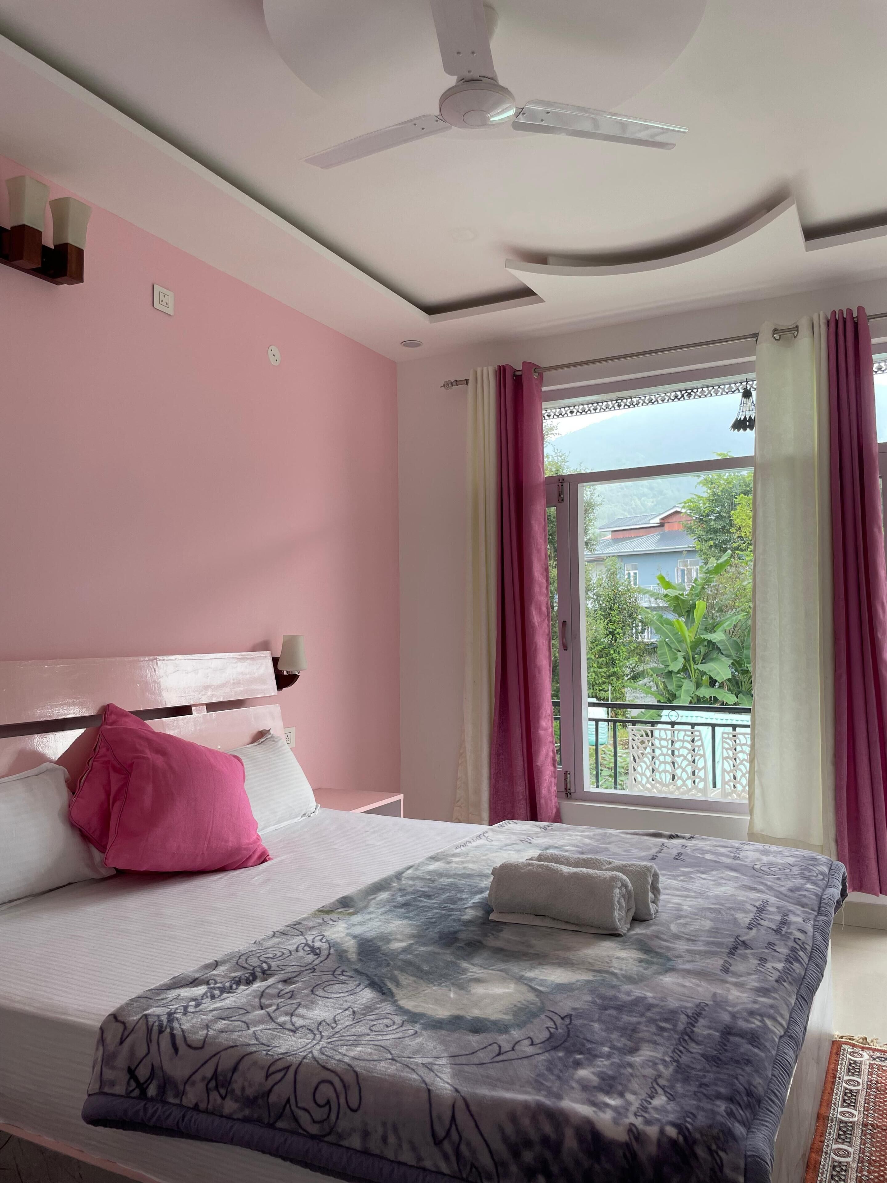 Un dormitorio con una cama, ventilador de techo, ventana con cortinas y vistas al exterior.
