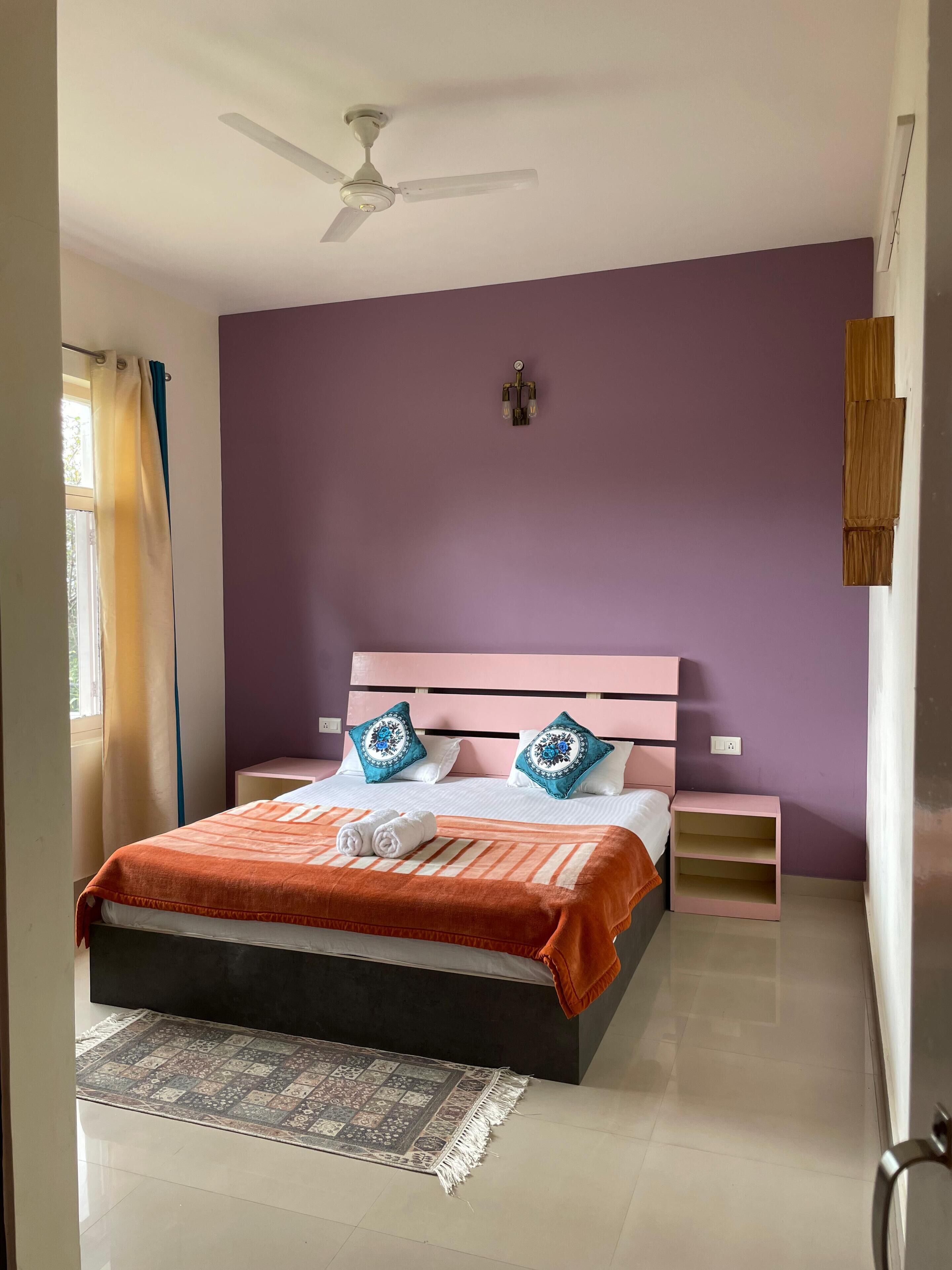 Un dormitorio con cama, ventilador de techo, mesita de noche y alfombra. Cuenta con una ventana con cortinas.