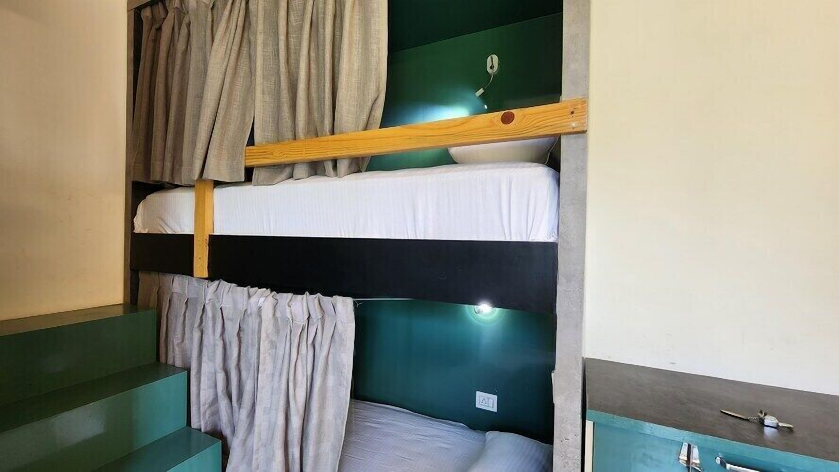 Una cama individual con estructura de madera, cabecera verde y colchón blanco. Hay un pequeño armario con tapa azul y base blanca.