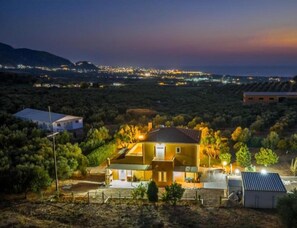 A house with a sloped roof, surrounded by vegetation and a distant cityscape at dusk. - Ktima Kika - Private Pool Escape Tranquility (Trifylia)