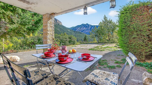 Un espace de restauration en plein air avec une table dressée pour deux personnes, un chien allongé à proximité et une chaîne de montagnes en arrière-plan.