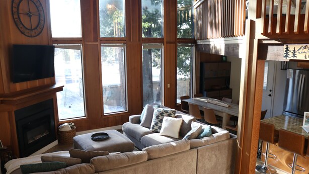 A spacious living room with a large sectional sofa, a fireplace, and a dining area with a high wooden ceiling and large windows.