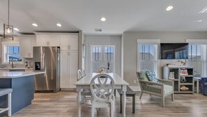 A modern kitchen with white cabinetry, a stainless steel refrigerator, and a dining area with a white table and chairs. - A Wave From It All.  Great location-short walk to beach and Kure Beach Pier. (Kure Beach)