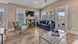 A spacious living room with a blue sofa, a wicker chair, a glass coffee table, and a flat-screen TV mounted on the wall. - A Wave From It All.  Great location-short walk to beach and Kure Beach Pier. (Kure Beach)