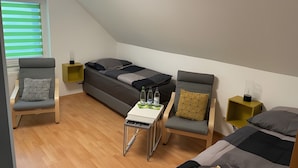 A modern, compact room with a loft bed, two chairs, a small table, and a window with blinds.
