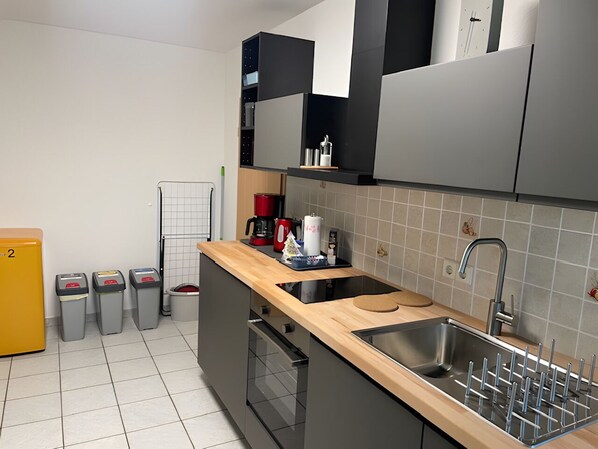 A modern kitchen with a wooden countertop, stainless steel sink, and grey cabinets. There are trash bins, a coffee maker, and a small plant on the counter.
