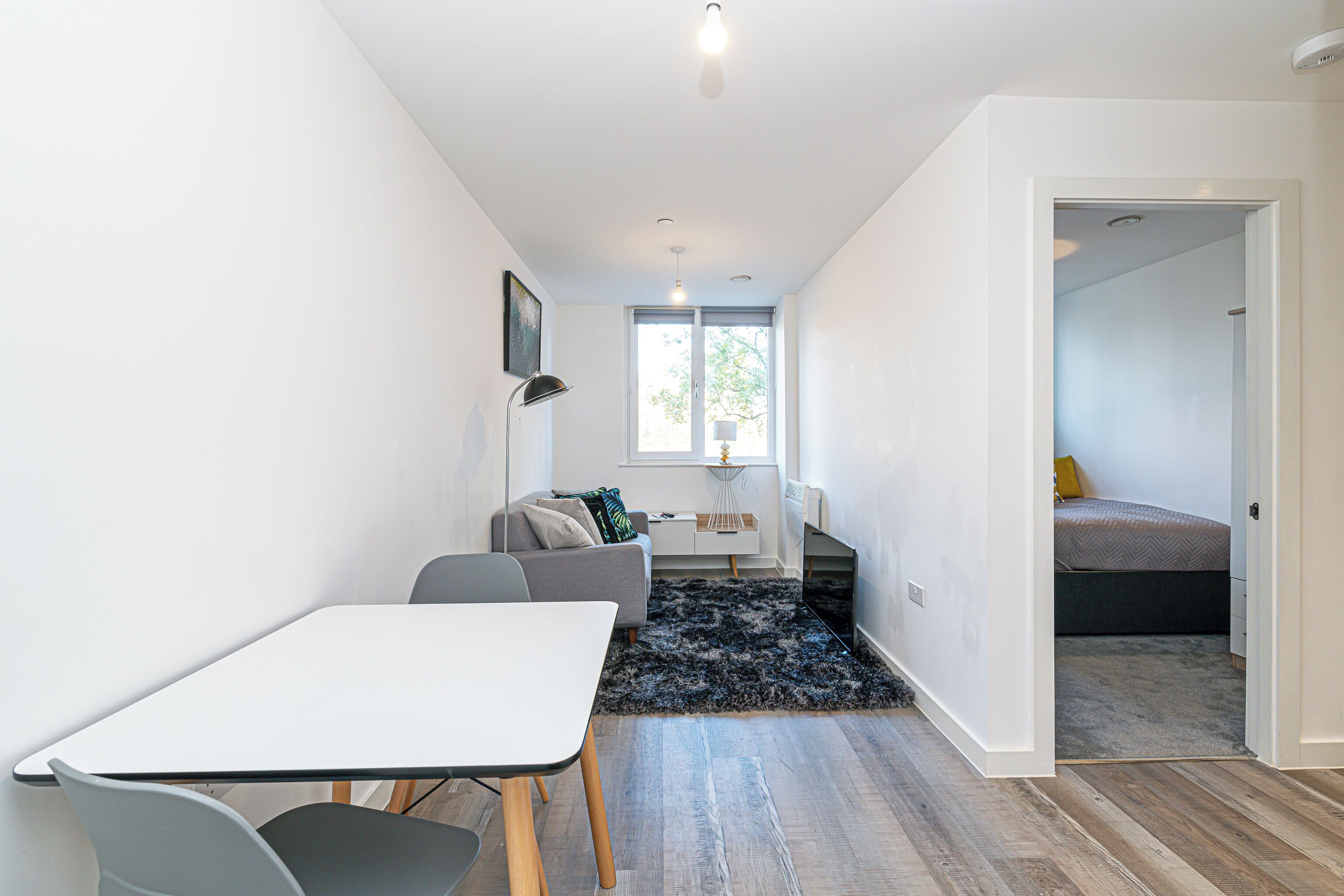 Un espacio habitable moderno y funcional con comedor, sofá, televisor y un dormitorio visible a través de una puerta abierta.
