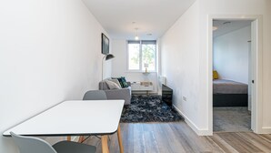 Ein moderner, kompakter Wohnraum mit Essbereich, einer Couch, einem Fernseher und einem durchgehenden Gang in den Schlafbereich.