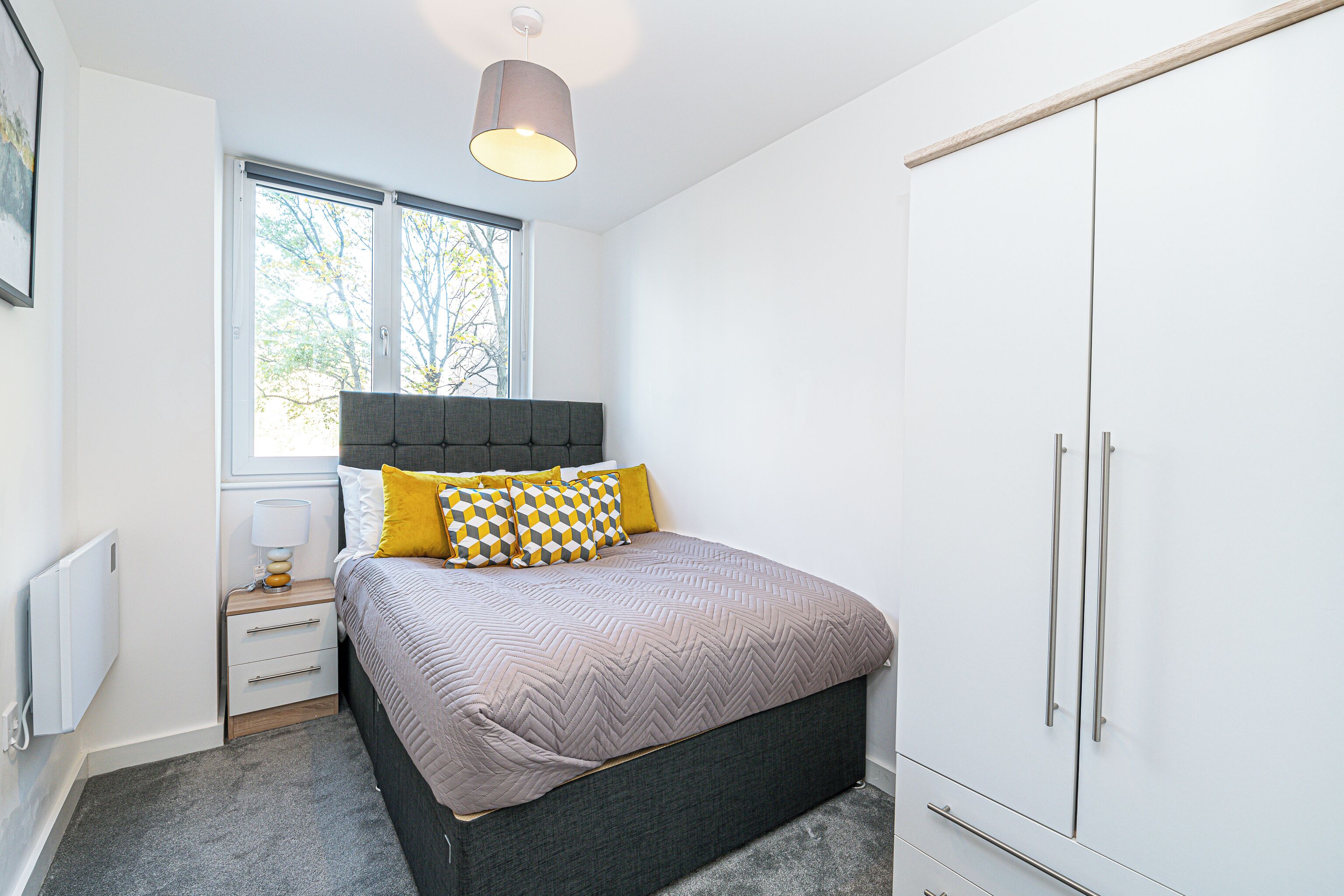Une chambre à coucher comprenant un lit, une table de chevet, une lampe, une armoire et une fenêtre donnant sur des arbres.