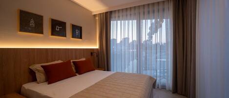 A hotel room with a large bed, a nightstand, and a city view through sheer curtains.