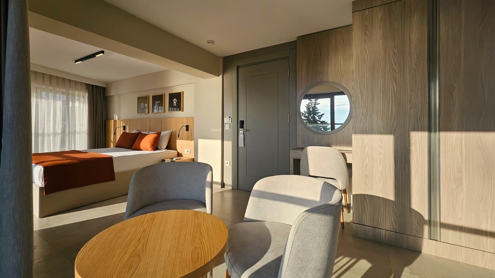 Ein modernes Hotelzimmer mit einem Bett, zwei Sesseln, einem runden Holztisch und einem großen Spiegel.
