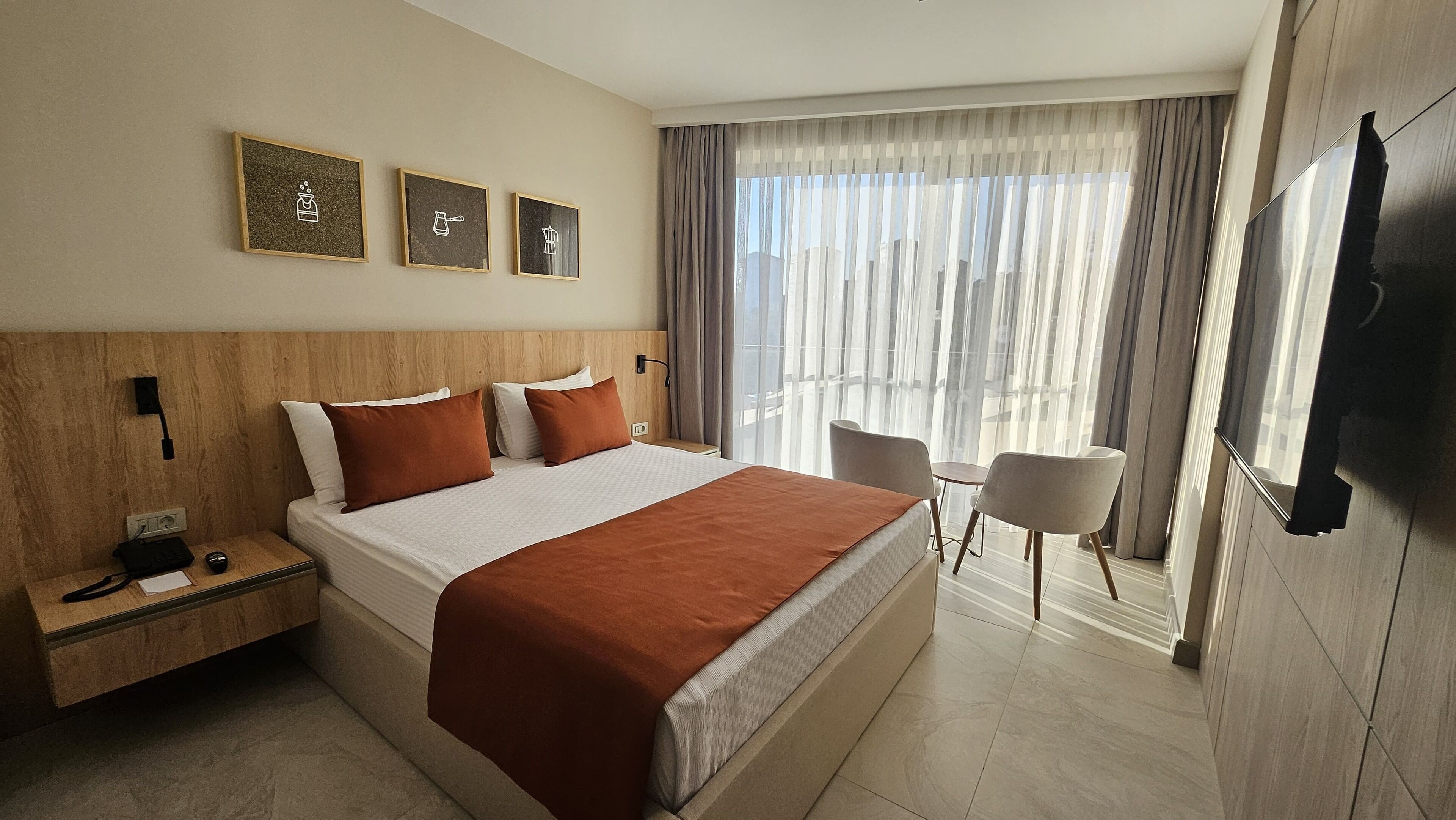 Habitación de hotel con una cama grande, una mesita de noche con teléfono y cargador, una pequeña mesa con silla, y un ventanal con cortinas.