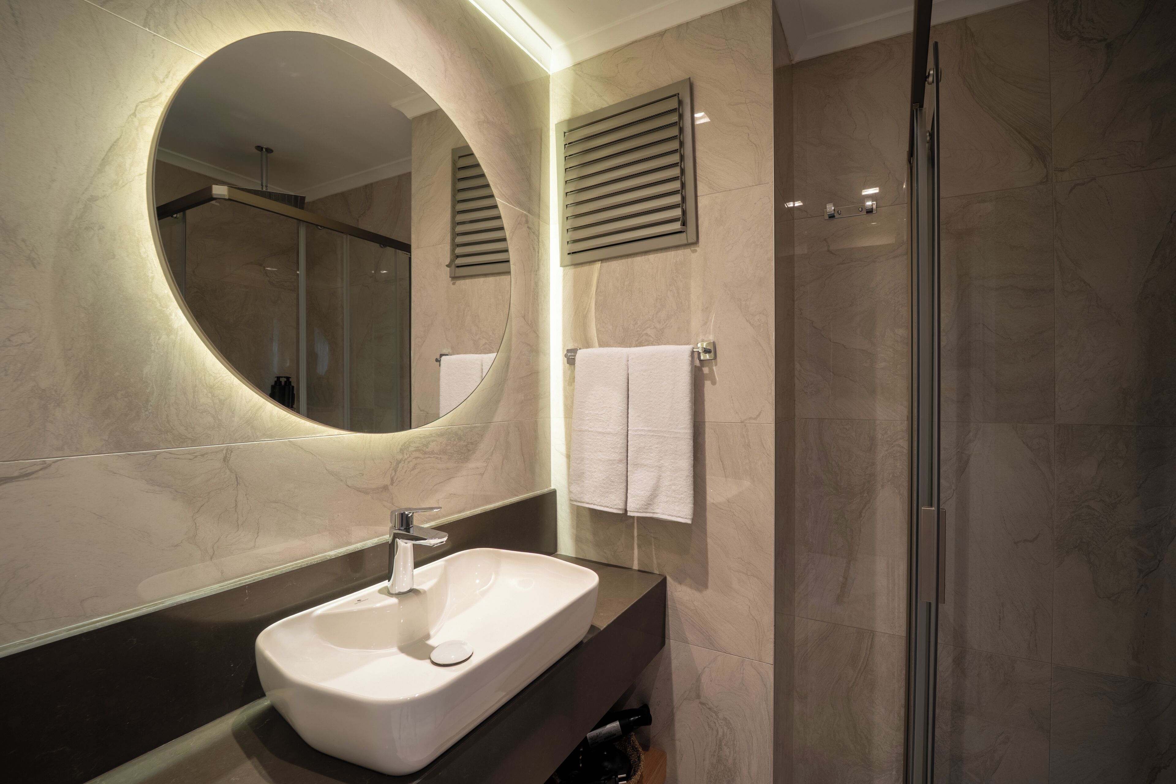 A modern bathroom with a large oval mirror, a white sink, and a glass shower enclosure.