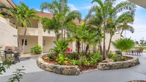Un patio con zona de barbacoa, mobiliario exterior y diversas plantas tropicales.