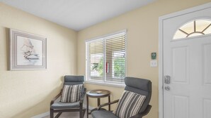 Una habitación con dos sillones grises, una mesita redonda pequeña, un cuadro enmarcado en la pared, una ventana con persianas y una puerta blanca.