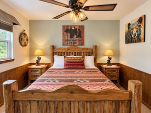 A wooden bed with a headboard, two bedside lamps, a ceiling fan, and a wall with a framed artwork. - Luxury Mountain Cabin w/ Hot Tub & Fireplace (Ruidoso)