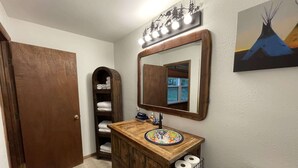 A bathroom with a wooden vanity, a mirror with lights, a towel rack, and a painting of a teepee. - Constancio's Cabin (Ruidoso)