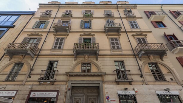 Un bâtiment historique doté de balcons richement décorés, d’une entrée centrale avec une porte ornée et de vitrines au rez-de-chaussée.