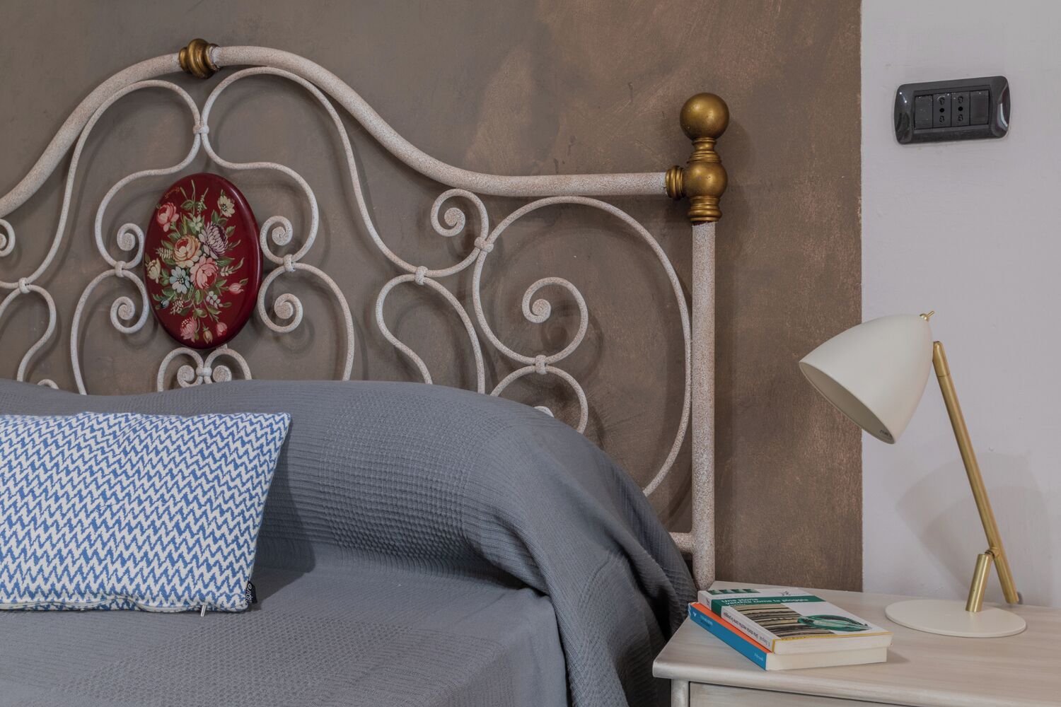 Un lit bien fait, avec une couverture grise et un coussin à motifs, une table de chevet avec des livres et une lampe, et un élément décoratif mural.