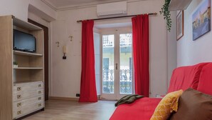 Une pièce avec un canapé rouge, un téléviseur posé sur une étagère en bois, un balcon avec des rideaux rouges et une plante.