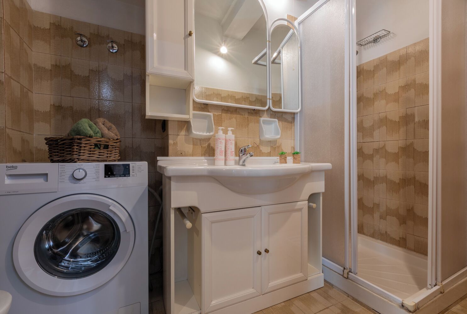 Un baño compacto con lavadora, lavabo con artículos de aseo y zona de ducha.