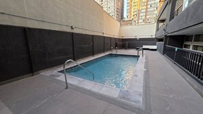 An indoor swimming pool with a metal ladder, tiled floor, and a view of high-rise buildings.