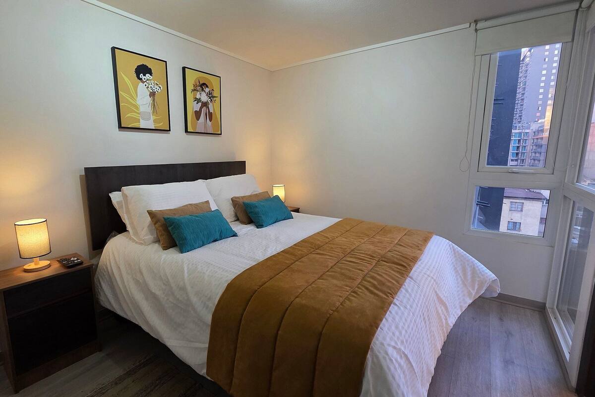 A neatly made bed with a brown blanket, white pillows, and blue accent pillows. There are two bedside lamps, a small nightstand, and two framed pictures on the wall.