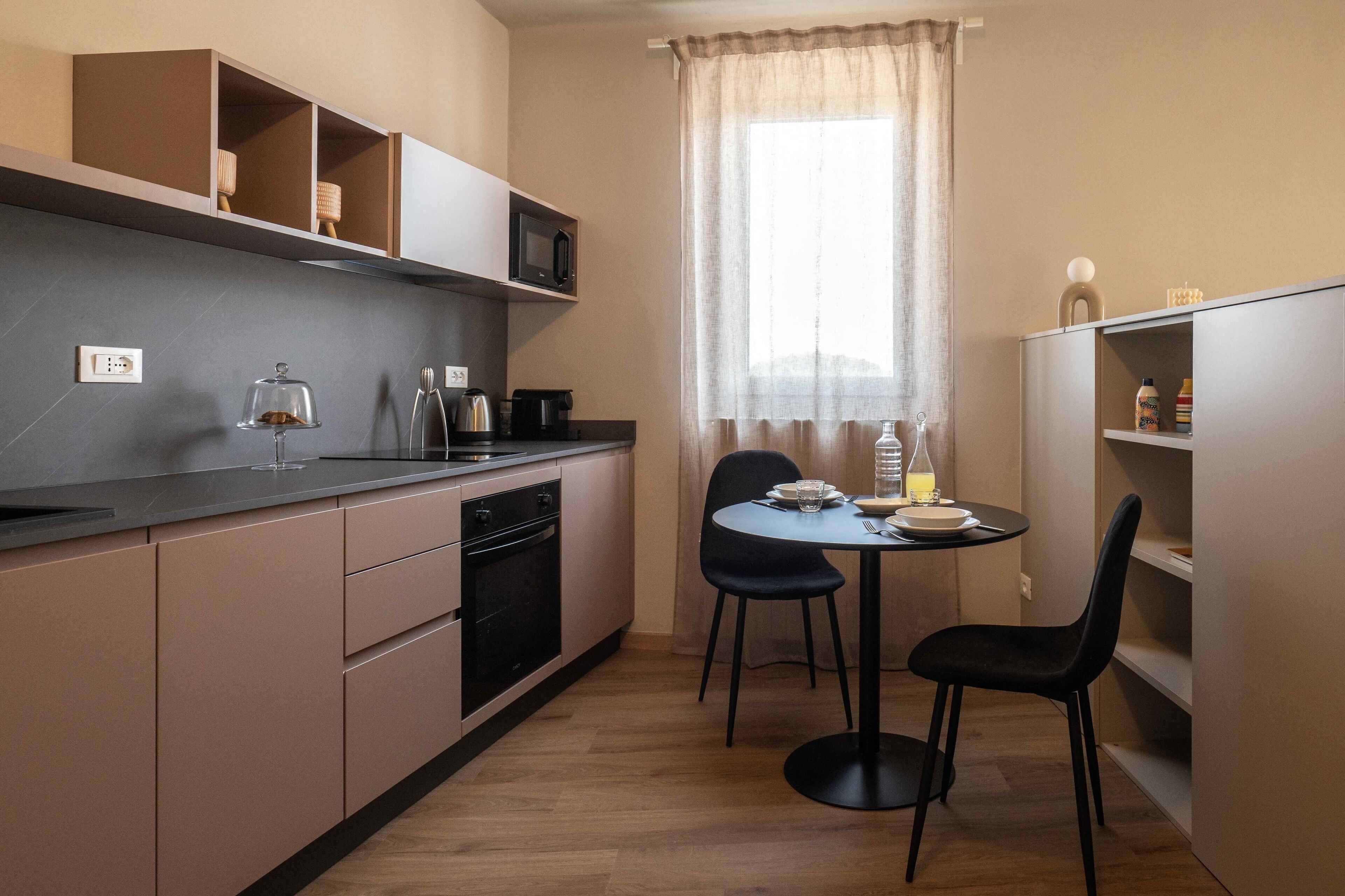 Une cuisine compacte équipée d’un micro-ondes, d’une bouilloire et d’une machine à café sur le plan de travail, d’une table à manger avec des chaises et d’une fenêtre avec des rideaux.