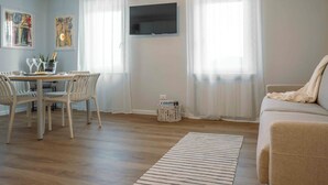 A living room with a sofa, a dining table with chairs, a flat-screen TV mounted on the wall, and a window with sheer curtains. - La Croce Apartments (Castelnovo ne' Monti)