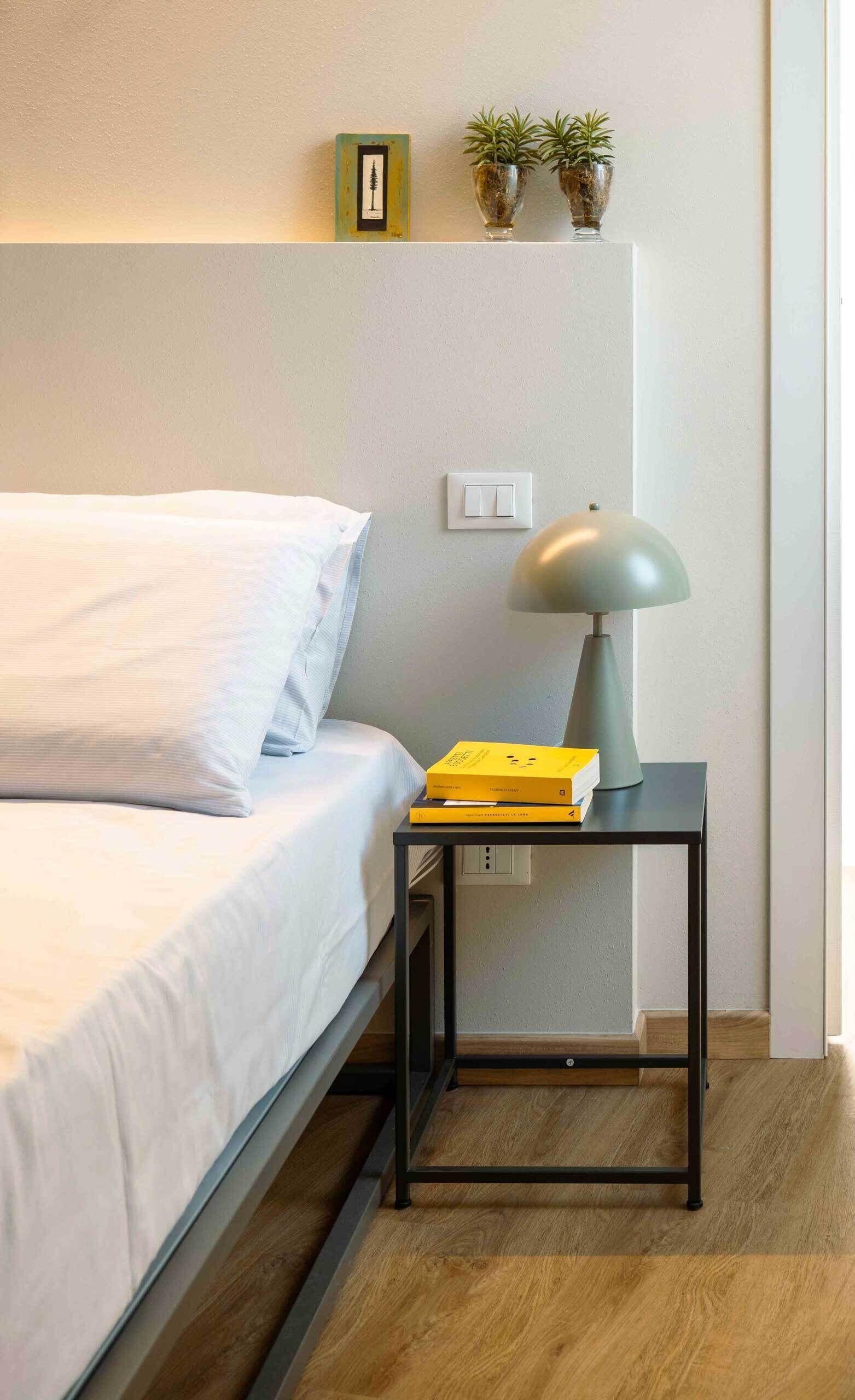 Un lit bien fait, avec du linge de lit blanc, une table de chevet avec une lampe, et deux plantes en pot accrochées au mur.