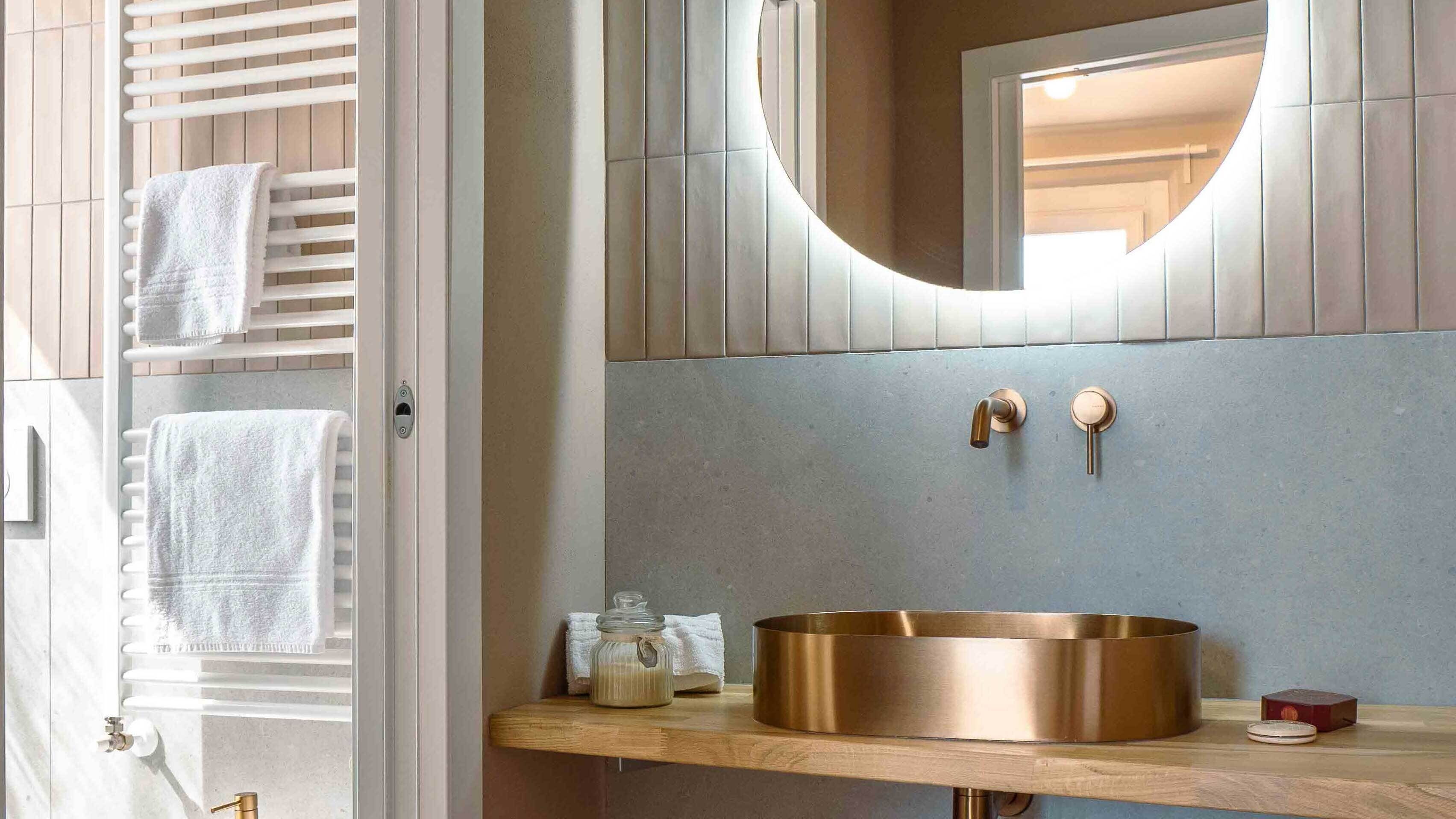 A bathroom with a round mirror, a copper sink, and a towel rack with white towels.