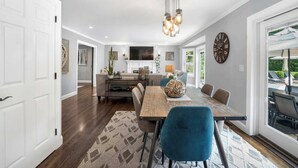 A spacious dining area with a wooden table and blue chairs, a chandelier, and a clock on the wall. - Southampton 4BR Home | Heated Pool, Firepit, Large Backyard (Hamptons)