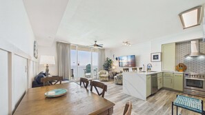 A modern kitchen with a central island, a dining area with a wooden table, and a living space with a sofa and a view of the ocean.