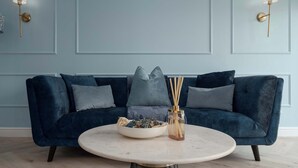 A blue velvet sofa with grey cushions, a round marble table with a glass vase and decorative items, and wall-mounted lamps.