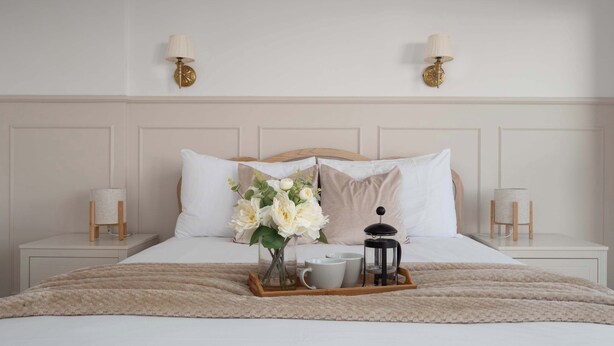 A neatly made bed with white linens, a fluffy beige blanket, and a bedside table with a lamp and a small vase with flowers.