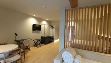 Une chambre d’hôtel moderne avec un plancher en bois, un canapé agrémenté de serviettes roulées, un bureau avec une chaise, un téléviseur fixé au mur et une cloison en bois.