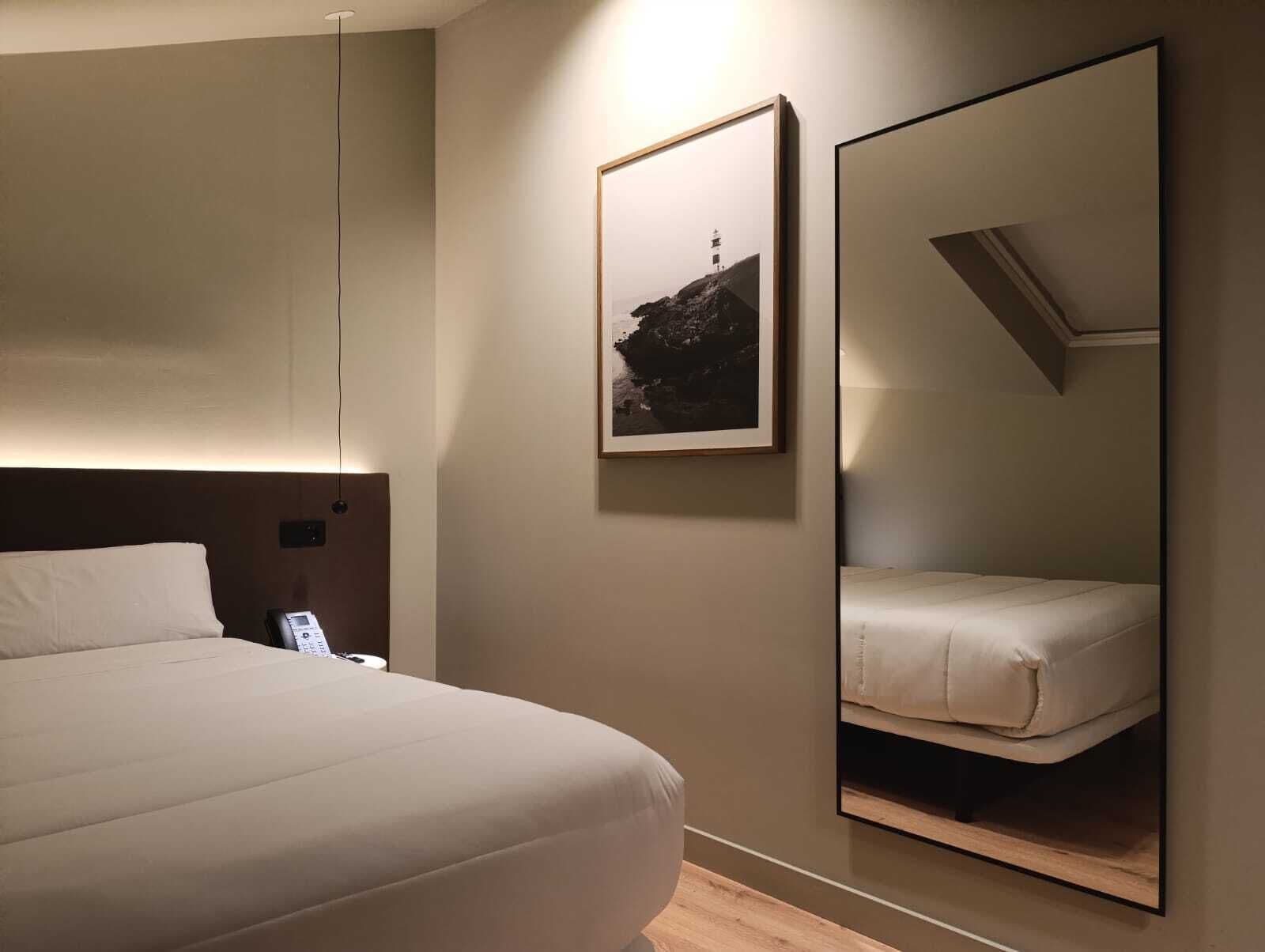 Habitación de hotel con una cama grande, una mesita de noche con un teléfono, un cuadro en la pared y un espejo grande que refleja la cama.